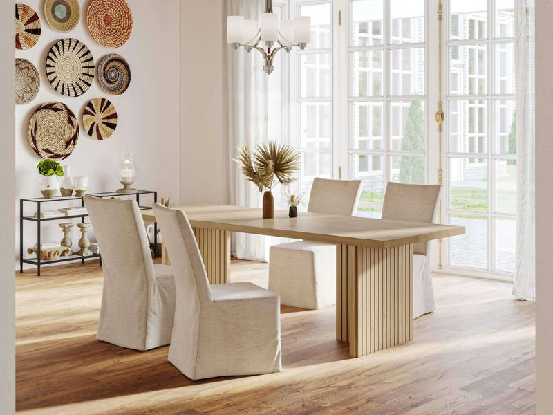 A modern dining room featuring the Francesca Dining Table in Satin from James+James, accompanied by four cushioned chairs with white slipcovers. Large windows allow natural light to flood the space. A wall showcases decorative woven baskets, and a sideboard holds an array of decorative items.
