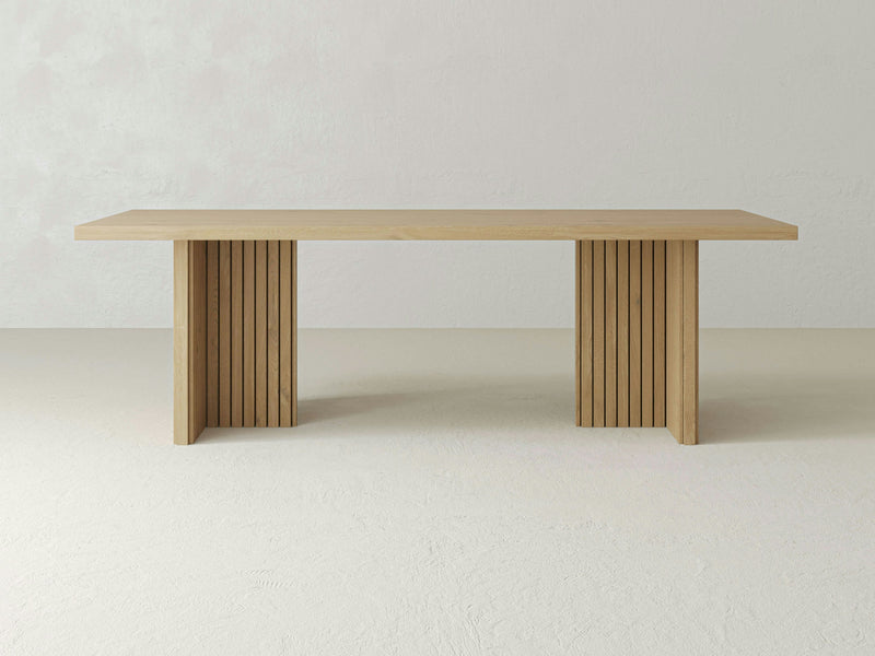 The Francesca Dining Table - Satin by James+James is centered in the image. This minimalist table features a rectangular top and two sturdy legs composed of vertical slats, creating a modern, sleek aesthetic. The background is light and plain, highlighting the table's clean lines and natural wood finish.