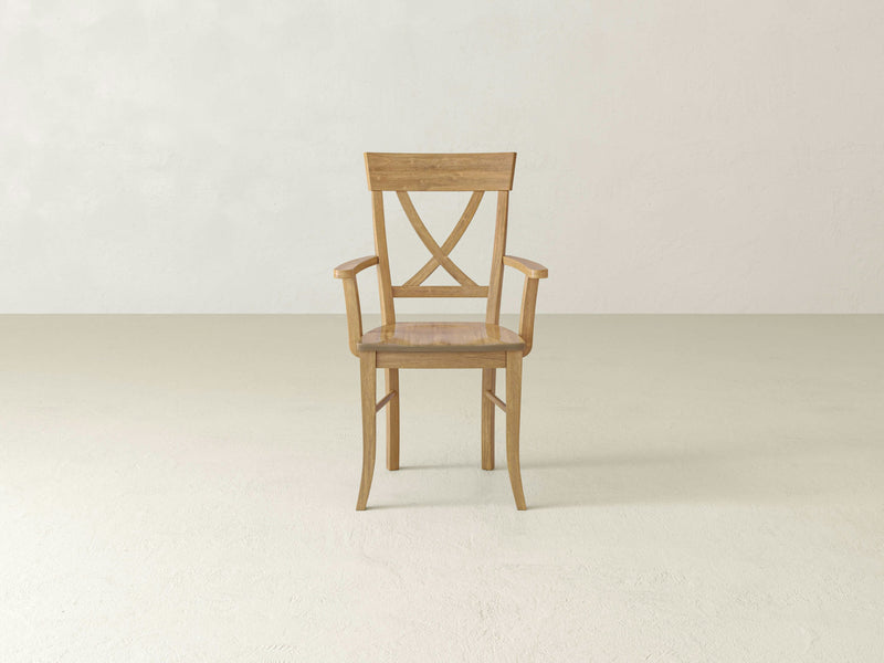 The Juniper Arm Dining Chair - Harvest Wheat from James+James is centered in the image, showcasing its simple, classic design. It features an X-shaped backrest, straight legs, and armrests. The background consists of a plain, light-colored wall and floor, creating a minimalistic and clean setting.