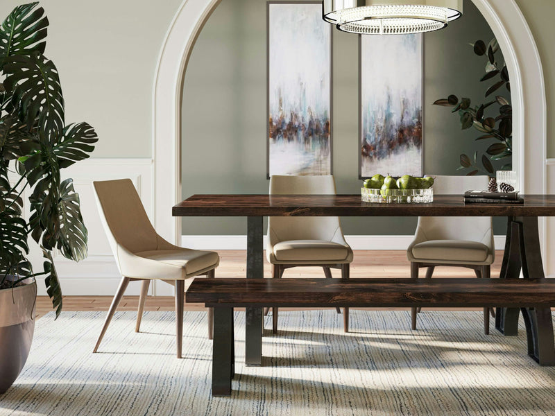 A modern dining room features a wooden table with the Wishbone Bench in Tobacco by James+James on one side and three beige chairs around it. A bowl of green apples sits on the table. Two abstract paintings hang on a light gray wall framed by a large arch, and a potted plant is in the foreground.