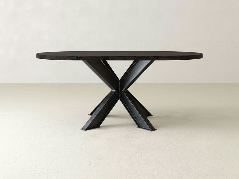 A sleek and modern dining table, the Shiloh Oval Dining Table by James+James features a deep grey wooden top and a distinctive X-shaped black metal base. Its minimalist design is complemented by the plain, light-colored wall and floor in the background.