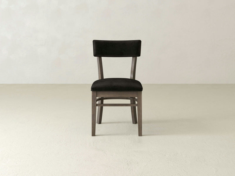 The Everly Side Dining Chair by James+James is a simple, modern piece featuring a dark upholstered seat and backrest that exudes timeless elegance. The chair boasts a wooden frame with four sturdy legs, perfectly placed in a minimalistic, light-colored room with a plain background.