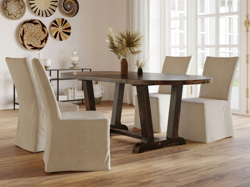 A modern dining room features the Thaden Oval Dining Table in tobacco finish from James+James, surrounded by four beige, slip-covered chairs on a wooden floor. The table is adorned with a brown vase holding dried plants. In the background, there's a sideboard displaying decorative items and woven wall art.