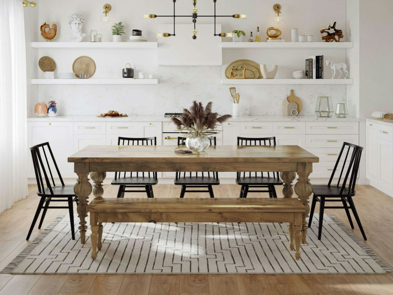 A cozy dining area featuring the Bailey Dining Table in Harvest Wheat by James+James, set for six with four black chairs and a wooden bench. The table holds a vase with dried flowers. In the background, a white kitchen with open shelving displays various decorations and kitchenware.