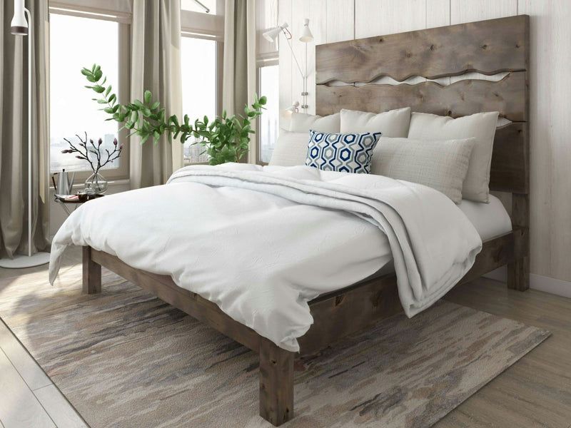 A cozy bedroom featuring the James+James Live Edge Bed - Barn Wood, adorned with white bedding and blue patterned accent pillows. Large windows with light-colored curtains allow abundant natural light to filter in. A potted plant by the window adds a touch of greenery to the room.