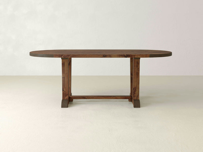 The Thaden Oval Dining Table - Tuscany by James+James showcases an excellently crafted wooden dining table with an oval-shaped top and a sturdy base featuring a rectangular support structure. Set against a plain, light-colored wall and floor, the table's craftsmanship and natural wood finish are prominently emphasized.