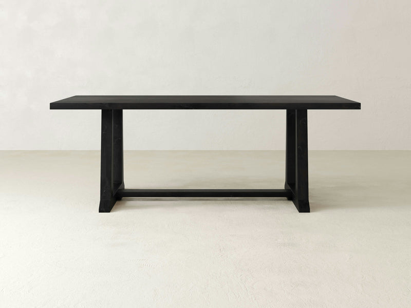 The Cora Dining Table - Charred Ember by James+James is a minimalist, rectangular black wooden dining table showcasing a simple yet sturdy design. It features two wide, flat legs connected by a horizontal support beam near the base and is set against a plain, light-colored background.