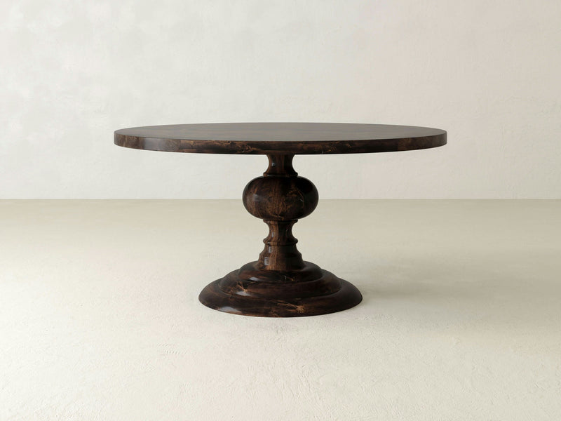 The Josephine Round Dining Table by James+James features a dark brown, round wooden surface with a smooth, polished finish and an ornate, turned pedestal base. Set against a light, plain backdrop, its elegant design and solid hardwood construction are beautifully highlighted.