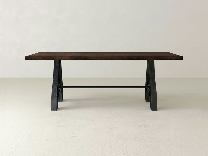 The Wishbone Dining Table - Tobacco by James+James is a rectangular wooden table with a rich dark finish and simple, clean lines. It features a thick, flat top and black metal legs in an angled A-frame design, connected by a horizontal metal bar at the base. The table stands on a light-colored floor against a plain background.