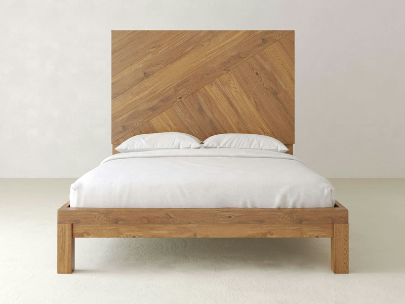 A Luna - Harvest Wheat wooden bed frame by James+James with a geometric headboard, showcasing a diagonal pattern of wooden slats. The bed is neatly made with white bedding and two white pillows placed against the headboard. The background features a plain, light-colored wall and floor.