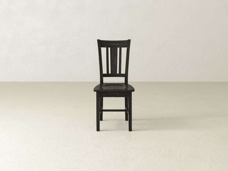 A William Dining Chair - Charred Ember, crafted by James+James, is centered on a plain, light-colored floor. The wooden chair features a slatted backrest and a flat seat with a simple, dark finish and no additional cushions or adornments. The background is a blank, off-white wall.