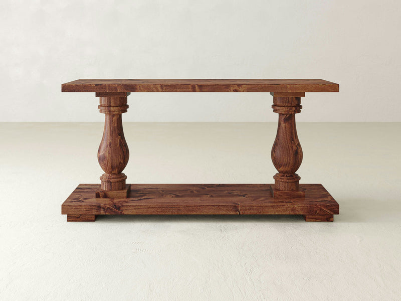 The Vivien Sofa Table - Tuscany by James+James is a wooden console table with a rustic finish, featuring a rectangular top and base connected by two ornately carved, spindle-like legs. The design is simple yet elegant, highlighting the natural wood grain.