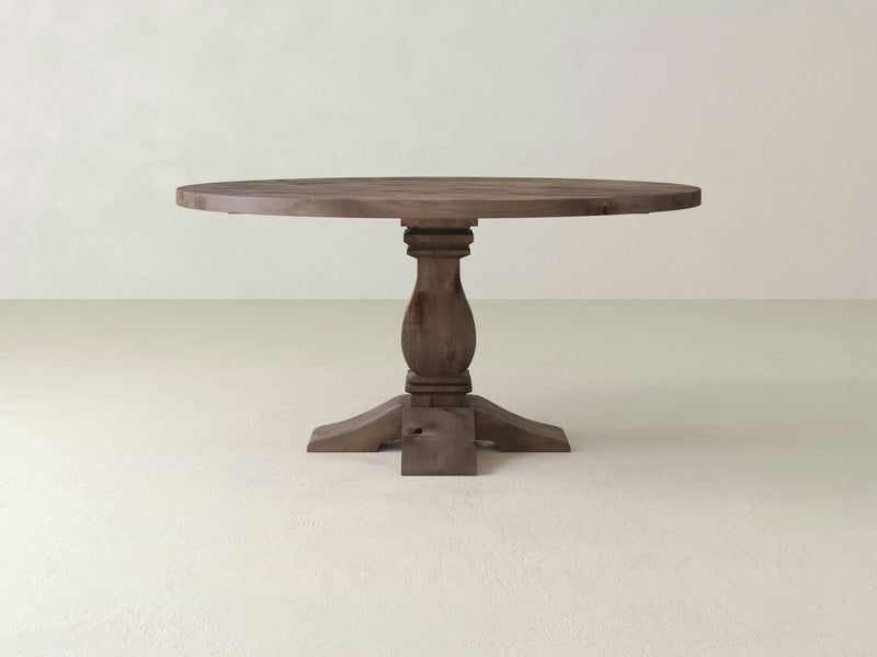 A Heirloom Round Dining Table - Barn Wood by James+James, featuring a smooth top and a single ornate pedestal base, is centered against a plain, light-colored background. The table's dark wood finish creates a striking contrast with the light floor and wall.