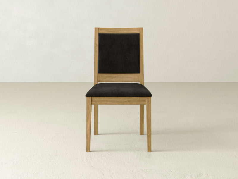 The Gina Side Dining Chair - Harvest Wheat by James+James, featuring a wooden frame with a black padded seat and backrest, sits on a light-colored floor against a plain, light-colored wall. The chair boasts a simple, modern design with straight lines and no armrests.