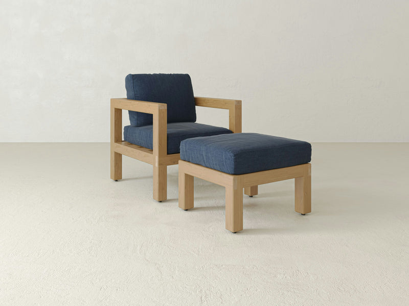 Introducing the Aria Outdoor Chair and Ottoman by James+James: a minimalist wooden outdoor chair featuring dark blue cushions on both the seat and backrest. Paired with a matching wooden ottoman topped with a dark blue cushion, these pieces are set against a pale, plain backdrop, perfect for enhancing your outdoor living space.