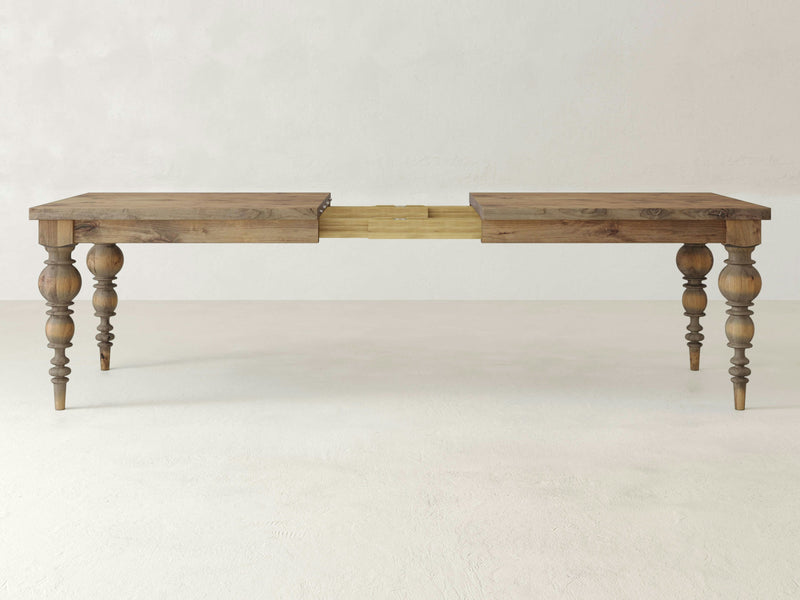 The Bailey Expandable Dining Table - Harvest Wheat by James+James is displayed with its ornate, turned legs and a central extension mechanism, ready to accommodate an additional leaf. The table features a rustic finish and stands on a plain, light-colored floor against a neutral background.