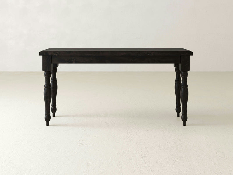 A James+James Abigail Sofa Table - Charred Ember with four ornate, carved legs stands alone on a light-colored floor against a bare, off-white wall. The tabletop has a simple rectangular shape, providing a contrast with the detailed craftsmanship of the legs.