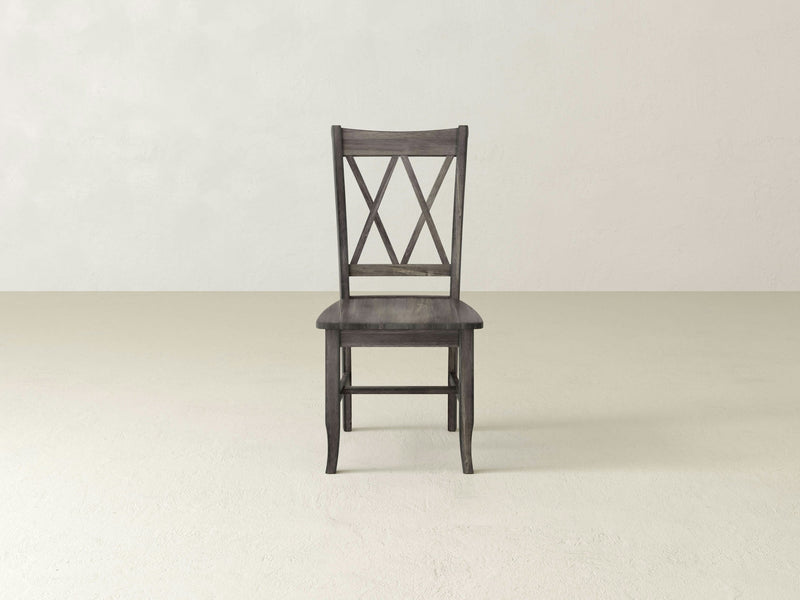 The James+James Double X-Back Dining Chair in Deep Grey is centered in an empty, light gray room. The chair features a straightforward design with a slightly curved seat and straight legs. The background is plain and unadorned.