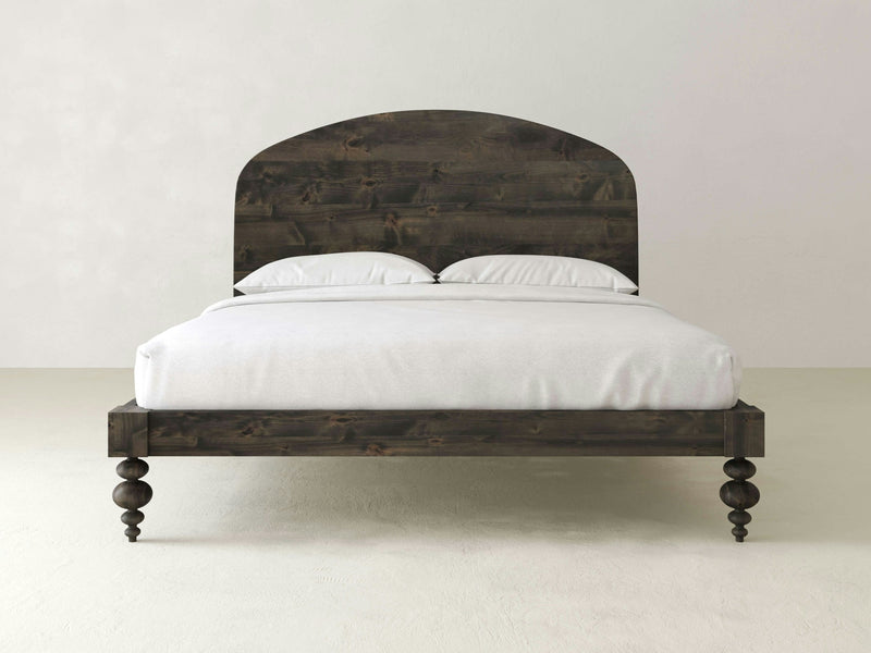 The Eden Bed in Deep Grey by James+James showcases a dark wooden bed frame with an arched headboard and turned legs, adorned with white bedding and two white pillows, set against a plain white background.