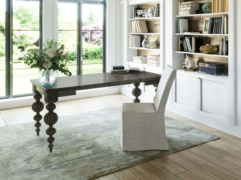 A bright home office with large windows overlooking a garden features the Olivia Desk in Deep Grey by James+James, characterized by its ornate legs. The space includes a light upholstered chair, a vase of flowers, and bookshelves adorned with various books and decorative items. The room is finished with a light area rug and wooden flooring.