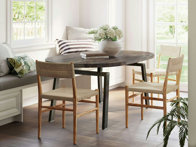 A cozy, sunlit dining nook features the Fulton Oval Dining Table in Tobacco by James+James, surrounded by three woven chairs. A cushioned window seat, adorned with striped and floral pillows, sits beside the table. A vase of white flowers is at the center, with large windows letting in natural light.