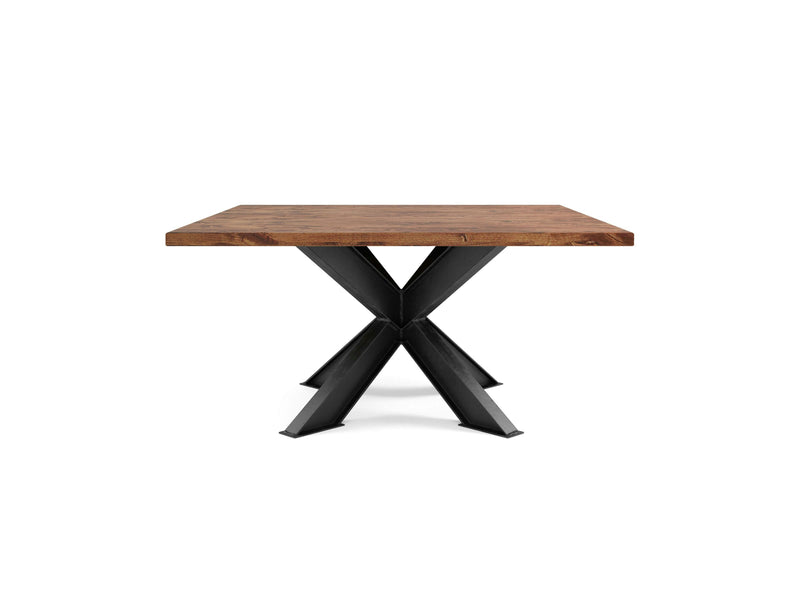 The Shiloh Square Dining Table - Tuscany by James+James is a modern and minimalist piece, featuring a square wooden top with a natural finish. It stands out with its unique black metal pedestal base, comprised of four angular legs converging at the center, emphasizing the striking contrast between the warm wood and industrial metal.