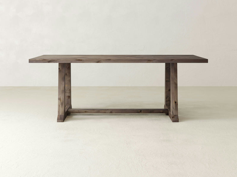 The Cora Dining Table - Barn Wood by James+James features a simple, rectangular design with a solid surface and sturdy legs connected by a horizontal brace. Placed against a plain, light-colored background, the table's natural finish showcases the beautiful grain and texture of the wood.