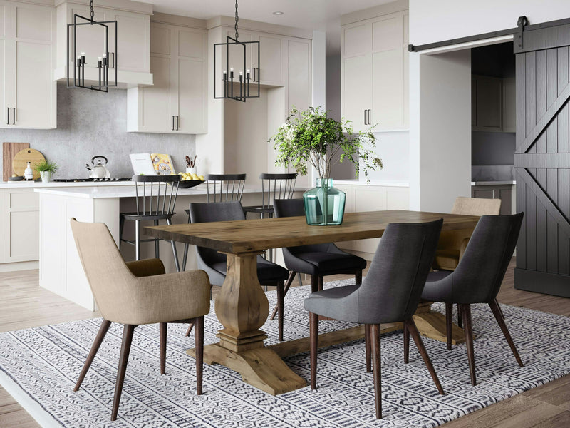 A modern, open-concept kitchen and dining area featuring light cabinetry, a kitchen island, and dark metal bar stools. The Heirloom Expandable Dining Table in Harvest Wheat by James+James stands at the center, surrounded by mixed fabric chairs on a patterned rug. Green plants in blue glass vases adorn the table. A sliding barn door is on the right.