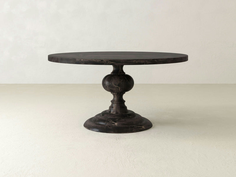 The Josephine Round Dining Table - Charred Ember from James+James is a round, dark-colored wooden table featuring a single pedestal base with an ornate design and a wide, sturdy bottom. It boasts a classic and elegant look, perfect for dining or decorative use against the plain white background.