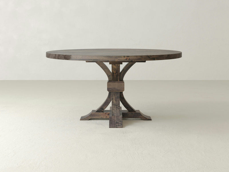 A round wooden table, known as the Violet Round Dining Table - Deep Grey by James+James, showcases a simple, rustic design. It stands on a single central pedestal with curved legs and features a natural wood grain finish. This elegant piece is set against a plain, light-colored background.