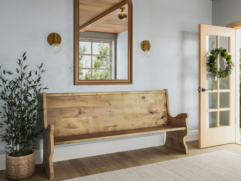 A minimalist entryway features the Rustic Church Pew Bench in Harvest Wheat by James+James, a tall potted plant, and a large mirror with a wooden frame. Two wall sconces with round bulbs flank the mirror. A door with glass panels is open, adorned with a green wreath, allowing natural light to stream in.