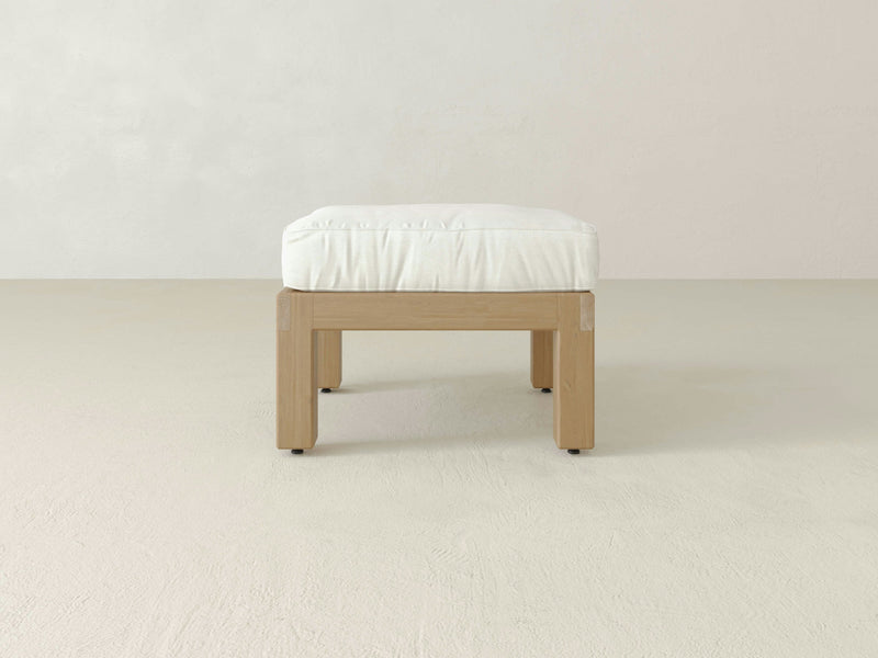 An Aria Outdoor Ottoman by James+James, featuring a handcrafted minimalist wooden frame paired with a white cushioned top, rests on a light-colored, smooth floor against an off-white wall. This ottoman showcases a clean, simple design with straight legs and an overall modern aesthetic, exuding subtle luxury.