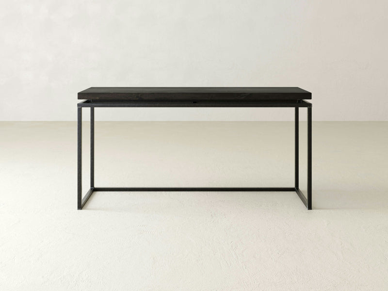 A minimalist Floating Top Sofa Table – Charred Ember by James+James stands in an empty, light-colored room with a plain wall and floor. The design is simple and modern, featuring clean lines and a smooth surface with a dark wooden top and black metal frame.