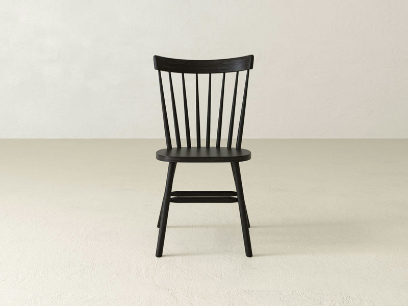 The Rustic Windsor Dining Chair - Charred Ember by James+James, a black wooden chair featuring a curved backrest with vertical slats and four slender legs, is shown at an angle. Its minimalist design evokes a classic, simple, and elegant look.