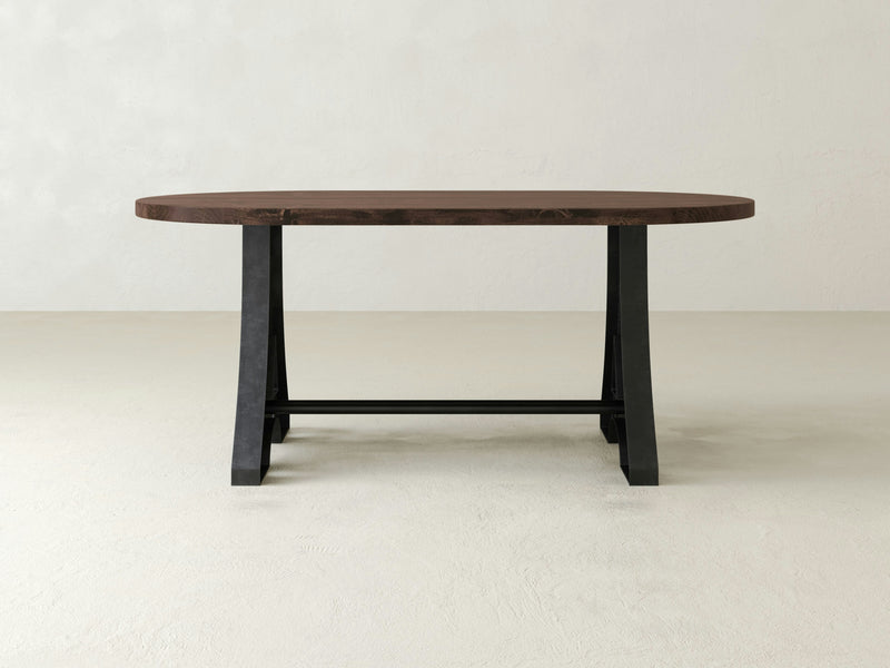 The Wishbone Oval Dining Table - Tobacco by James+James features a minimalist design with a dark brown wooden top and black metal legs, set against a plain off-white background. The simple design highlights the contrast between the rich wood grain of the tabletop and the sleek, industrial look of the metal base.