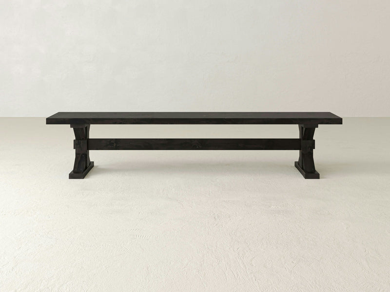 A long, rectangular bench named the Trestle Bench - Charred Ember from James+James features a minimalist design in charred black wood. The bench includes two sturdy X-shaped legs and is designed to seat multiple people, making it ideal for both outdoor and indoor settings. It is showcased against a plain white background.