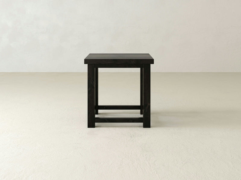 The Carter X Side Table - Charred Ember from James+James stands against a plain off-white backdrop. This minimalist black wooden piece features a rectangular top with simple, straight legs and a basic support structure, embodying a clean and modern design aesthetic.