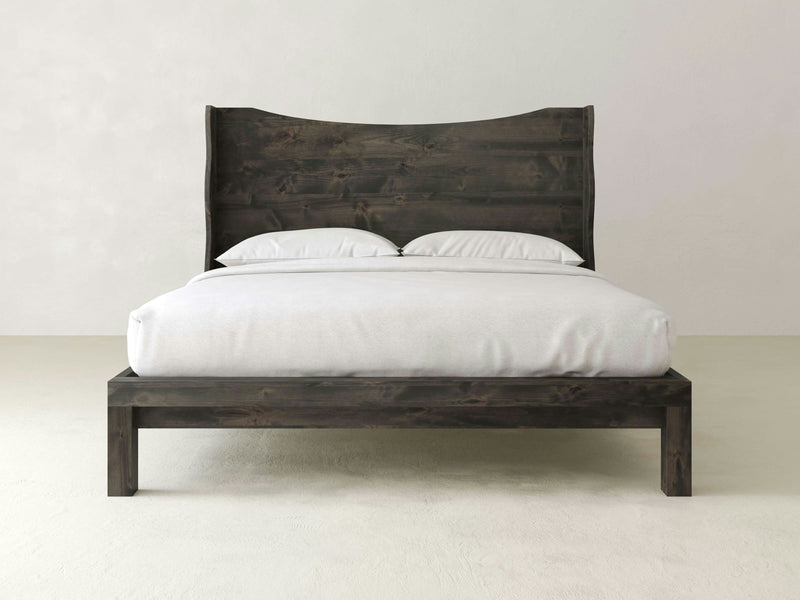 A minimalist wooden bed from James+James, named the Harlow Bed in Deep Grey, featuring a rustic finish and a tall, curved headboard. The bed is neatly made with a white fitted sheet, blanket, and two white pillows. The background is a plain, light-colored wall and floor.