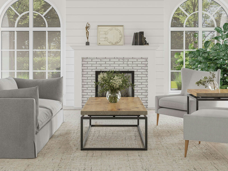 A cozy living room features a light gray sofa and armchairs around a James+James Floating Top Coffee Table in Harvest Wheat with a vase of flowers. In the background, there is a white brick fireplace with decor on the mantel and two large arched windows letting in natural light.