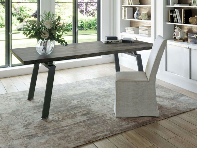 A contemporary home office features the stylish Fulton Desk in Deep Grey by James+James, positioned on a light-patterned rug. An upholstered chair is placed in front of the desk, which is adorned with a glass vase of white flowers, a book, and a stack of papers. In the background are shelves and a window.