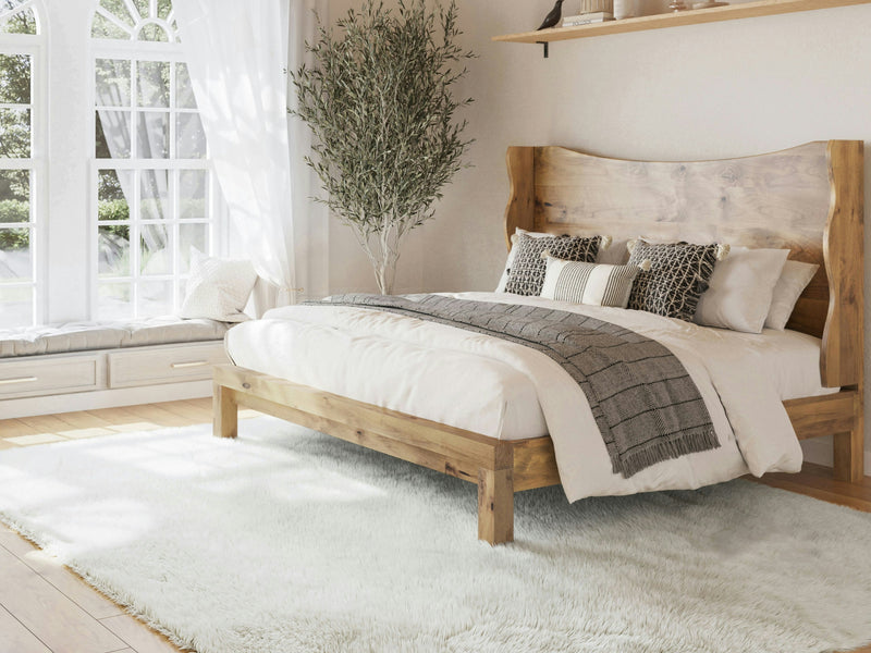 A cozy bedroom showcases the Harlow Bed in Harvest Wheat from James+James, adorned with elegant white and gray bedding, a complementary gray throw, and an array of decorative pillows. A tree stands beside the bed, which is set on a luxurious white rug. Sunlight pours through a large window with a cushioned seat, gently illuminating the space.