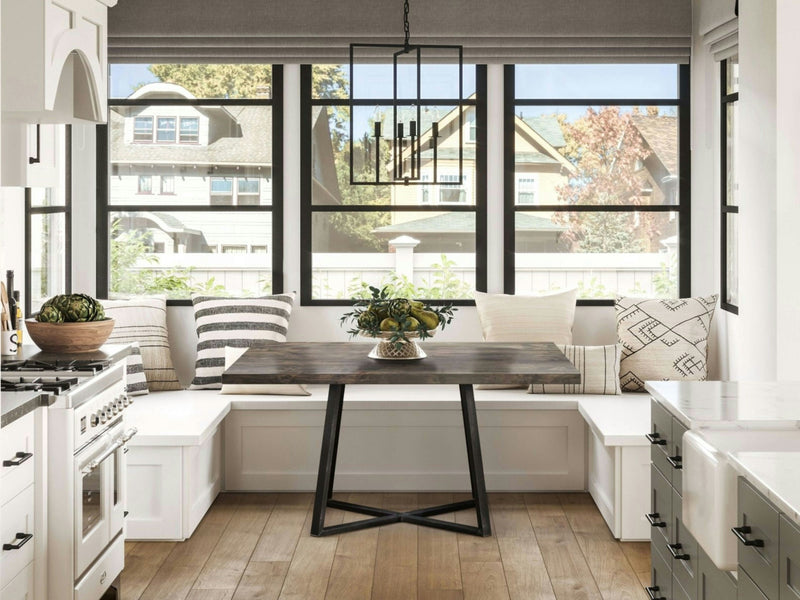 A modern kitchen with large windows featuring the Watson Square Dining Table in Deep Grey from James+James, complemented by black metal legs. The space also includes white built-in bench seating with patterned cushions, white cabinets, a visible gas stove, and pendant lighting above the table.