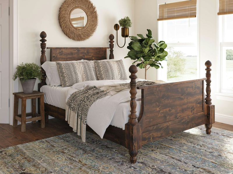 A cozy bedroom featuring the Catherine Bed in Tobacco by James+James, adorned with white and patterned bedding. A round mirror with a woven frame hangs above the bed. A wooden nightstand with a potted plant is on one side. Large windows with bamboo blinds allow natural light to fill the room.