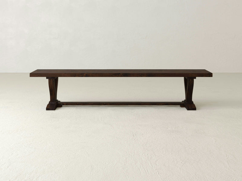 The Vera Bench - Tobacco by James+James features a dark wooden finish with a rectangular seat, characterized by its simple yet sturdy design. The bench sports slightly curved legs connected by a horizontal support bar and is showcased against a plain, light-colored background.