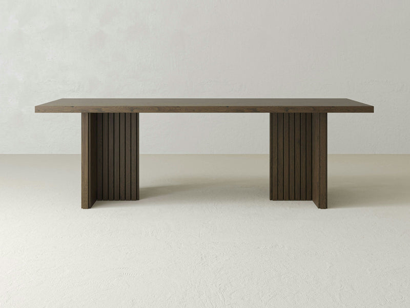 The Francesca Dining Table by James+James, crafted from white oak hardwood, showcases a minimalist rectangular top and two sturdy, ribbed panel legs. Set against a light-colored background, its Scandinavian-inspired design is highlighted in its sleek and modern form.