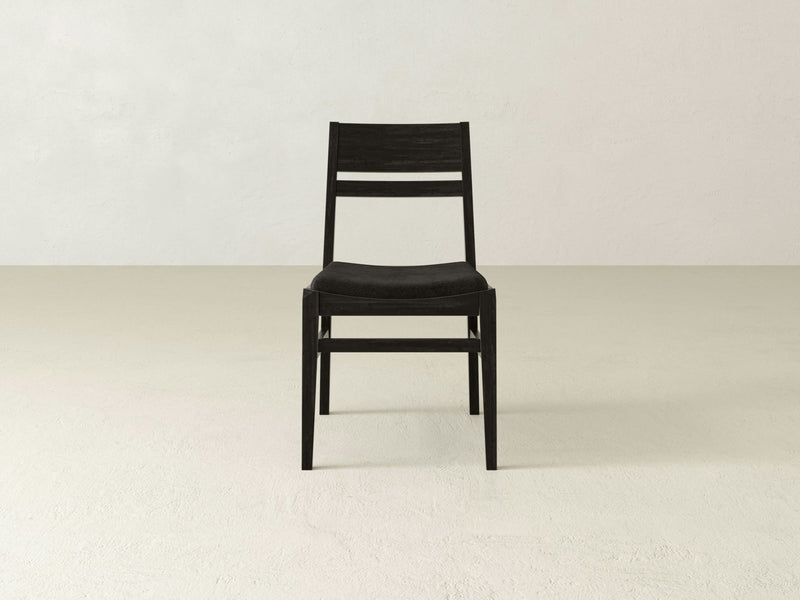 The Delilah Dining Chair by James+James, with its minimalist black design and wooden frame, sits on a neutral, cream-colored floor. The chair features a straightforward design with a straight back, thin horizontal slats on the backrest, and an upholstered seat. The light and plain background wall enhances the scene's clean, modern aesthetic.