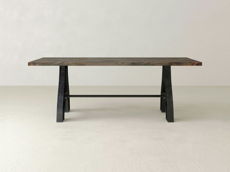 A rectangular Wishbone Dining Table - Deep Grey from James+James with a dark brown tabletop and black metal legs stands on a light gray floor against a light gray wall. The table features a modern, minimalist design with clean lines and a sturdy appearance.