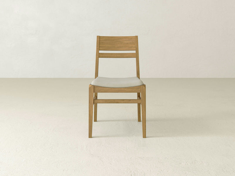 The Delilah Dining Chair - Harvest Wheat by James+James is a simple wooden chair with a light cushion seat, positioned in an empty room with plain light-colored walls and floor. The chair boasts a minimalist design, featuring a straight backrest with two horizontal slats.