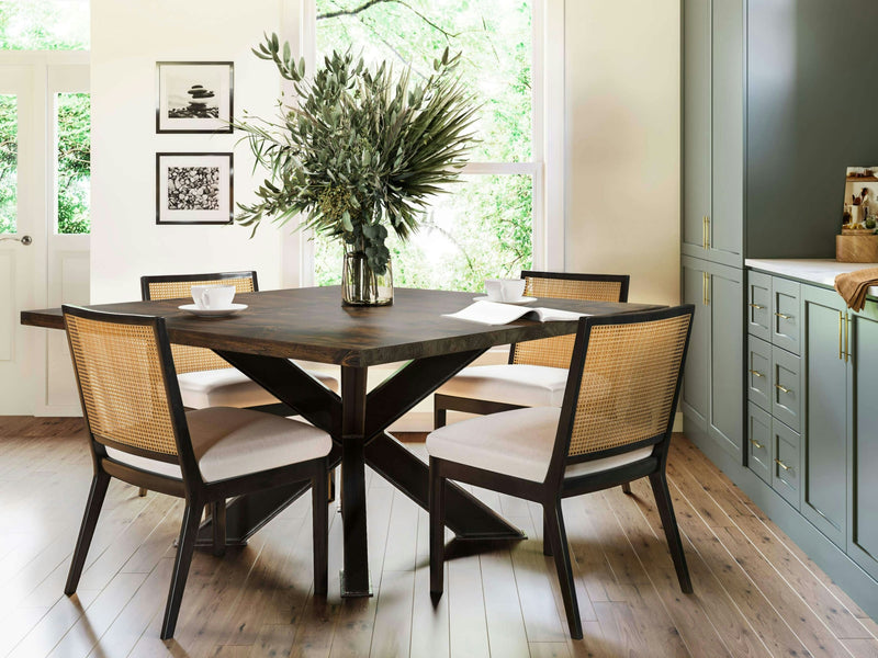 A modern dining room features the Shiloh Square Dining Table in Tobacco by James+James, accompanied by four wicker-backed chairs. A large vase with green foliage is placed at the center of the table. The room has light wood flooring, white walls adorned with framed art, and a green cabinet with drawers in the background. Natural light fills the space.