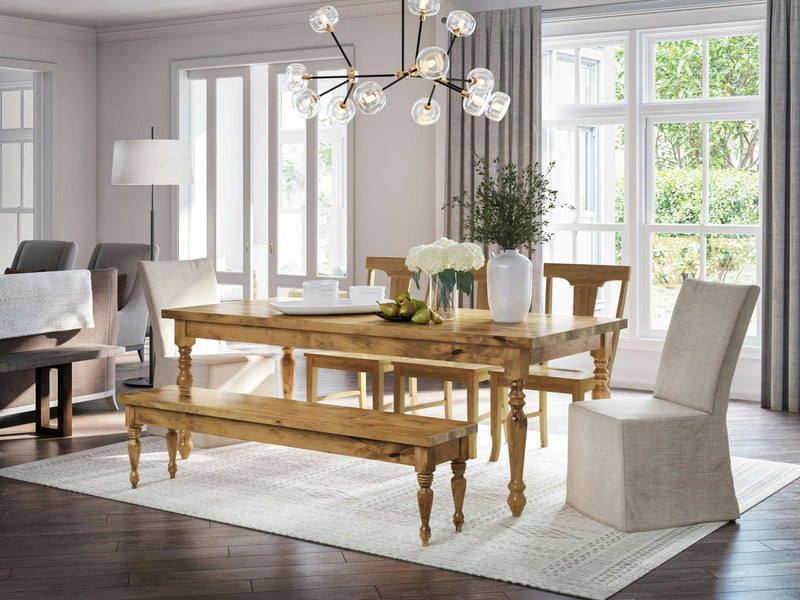 A bright dining room with the French Country Expandable Dining Table in Harvest Wheat by James+James, set for six. It features a bench on one side and cushioned chairs on the other sides. A modern chandelier hangs above the table, which is adorned with white flowers and a bowl of fruit. The room has large windows with beige curtains.